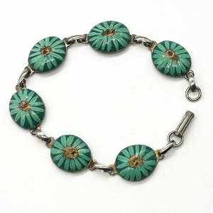 May include: A link bracelet featuring six oval-shaped charms, each decorated with a teal green daisy flower pattern with a textured gold-tone center. The charms are connected by silver-tone metal links and include a secure metal clasp closure.