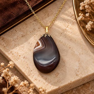 May include: A teardrop-shaped pendant necklace featuring a polished dark brown and white banded agate stone. The stone is secured by a gold-toned metal bail and hangs from a delicate gold-toned chain, showcasing natural swirling patterns.