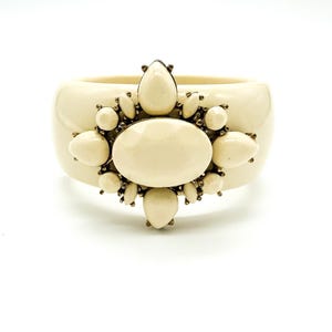 May include: A wide cream-coloured bangle bracelet featuring a central oval cabochon surrounded by smaller teardrop and round cream-coloured stones set in a dark metal frame. The smooth, polished surface of the bangle contrasts with the ornate cluster.