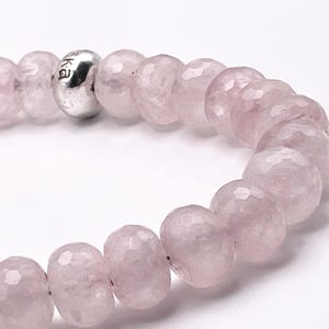 May include: A rose quartz beaded bracelet with a faceted design. The light pink beads are strung together with a silver-colored bead that has the letters "ka" engraved on it. The bracelet is on a white background.