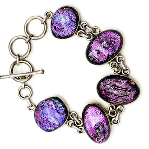 Op de afbeelding: Een zilverkleurige armband met vijf ovale en afgeronde glazen cabochons met een glinsterende paarse en iriserende afwerking. De schakels zijn verbonden door kleine zilveren ringen en het sieraad heeft een sluiting met een staafje.