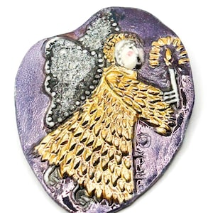 May include: A ceramic brooch featuring a stylized angel figure with gold-toned textured wings and robes, holding a candle. The figure is set against a textured, irregular purple background with a glossy finish and visible artistic detailing.