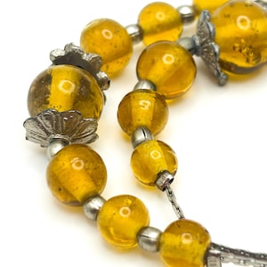 May include: Close-up of a necklace with large, translucent amber-coloured beads. The beads are interspersed with silver-toned metal accents and strung on a delicate chain. The necklace is set against a white background.