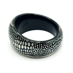 May include: A wide, circular bangle bracelet featuring a glossy black base with a repeating pattern of small, irregular silver-toned spots covering the exterior surface. The interior of the bracelet is smooth, solid black with a polished finish.