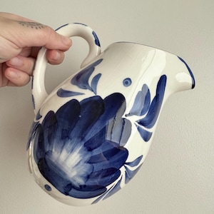 May include: White ceramic jug with a curved handle and spout. The jug is decorated with a large blue floral design and blue leaf accents. The jug is held by a hand.