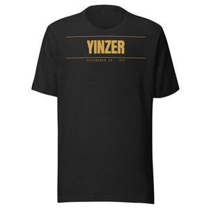 May include: A black short-sleeve t-shirt featuring the word YINZER in gold capital letters centered on the chest. Below the word, two thin horizontal gold lines frame the text PITTSBURGH, PA 412 in a smaller gold font, creating a clean graphic design.