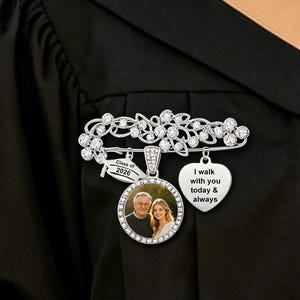 May include: A silver brooch with a floral design and clear crystals. The brooch has three pendants: a round pendant with a photo, a heart-shaped pendant with text, and a banner pendant with the text "Class of 2020". The brooch is pinned to a black graduation gown.