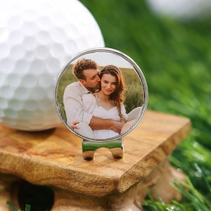 Peut inclure: Une balle de golf blanche est posée sur un support de tee en bois, à côté d'un marqueur de balle personnalisé. Le marqueur présente une photo d'un couple dans un cadre argenté. La scène se déroule sur une surface herbeuse verte.