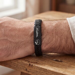May include: Black braided leather bracelet with a rectangular metal clasp. The clasp is engraved with the name "Alton" and a heart with a paw print. The bracelet is worn on a wrist.