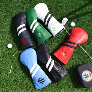 Könnte beinhalten: Eine Auswahl an Golfschlägerhauben in verschiedenen Farben, darunter Marineblau, Grün, Weiß, Rot, Schwarz und Hellblau. Jede Haube hat weiße Streifen und personalisierte Initialen oder Designs. Golfbälle und Tees liegen auf dem grünen Gras.