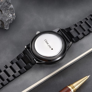 May include: A black wristwatch with a matching strap. The watch face is white and features the words "I love you" and a heart symbol. The watch is on a grey surface.