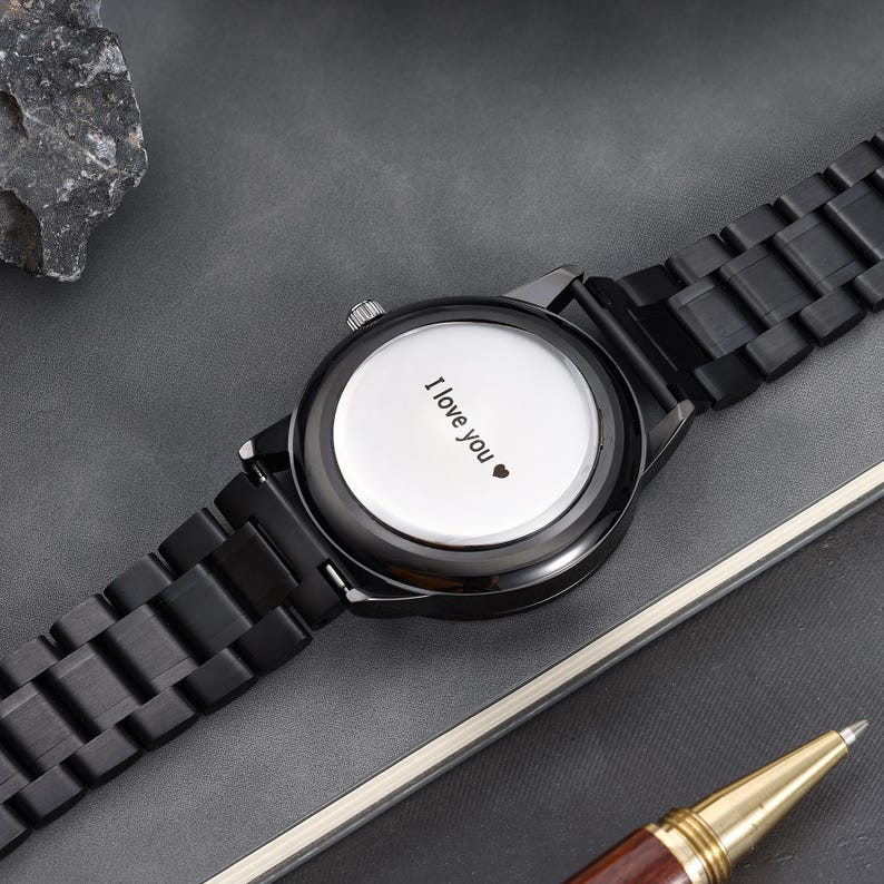 May include: A black wristwatch with a matching strap. The watch face is white and features the words "I love you" and a heart symbol. The watch is on a grey surface.