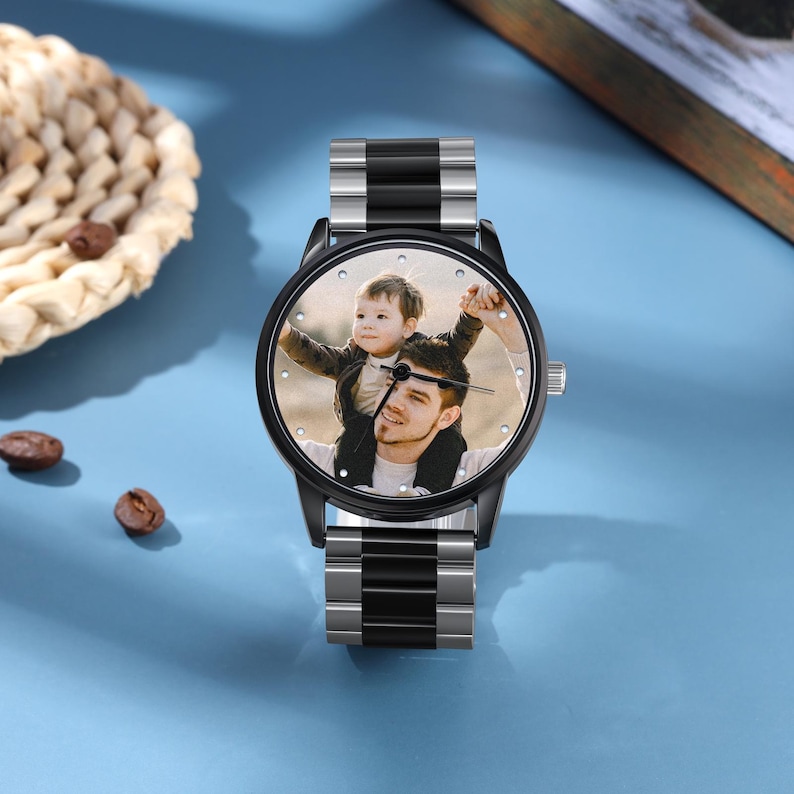 May include: A black and silver wristwatch featuring a photograph of a man and a child on the watch face. The watch has a round face and a strap with silver and black links. The background is a light blue surface.