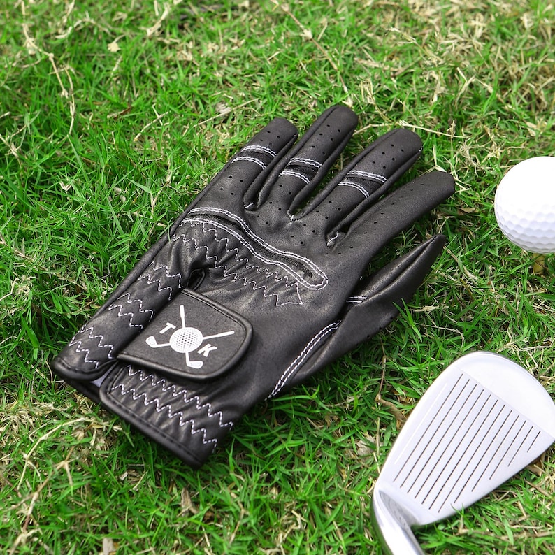 May include: Black golf glove with white stitching and a golf club logo, resting on green grass next to a golf ball and a golf club. The glove has small ventilation holes.