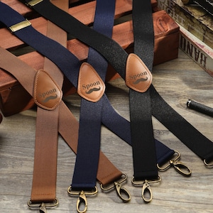 Peut inclure: Diverses bretelles marron, bleu marine et noires. Chacune est ornée d'un patch en cuir avec le mot "Spoon" et une moustache. Les bretelles ont des attaches et des ajusteurs en métal doré, présentées sur une surface en bois.