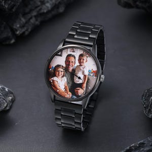 Pode incluir: Um relógio de pulso preto com uma pulseira de metal. O mostrador do relógio exibe uma fotografia colorida de um homem e duas crianças. O relógio está em um fundo escuro com pedras cinza escuro.