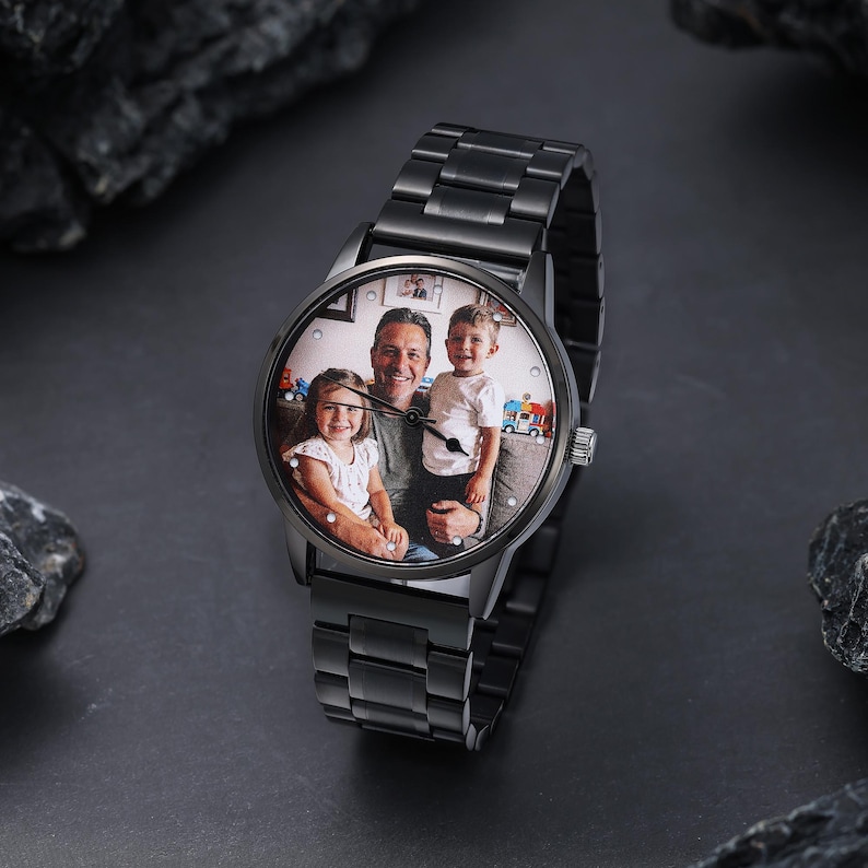 May include: A black wristwatch with a metal strap. The watch face features a colour photograph of a man and two children. The watch is set against a dark background with dark grey rocks.