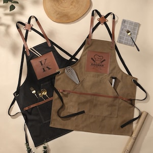 Może przedstawiać: Dwa fartuchy, jeden czarny i jeden brązowy, ze skórzanymi detalami i paskami. Czarny fartuch ma inicjały "IZ" i imię "Kevin" na skórzanej naszywce. Brązowy fartuch ma logo czapki kucharskiej i napis "DAGMAR". Oba mają kieszenie.