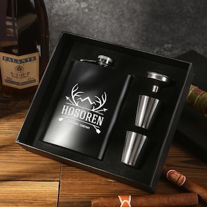 May include: A black flask gift set with the word "HOSOREN" in white, along with a mountain and antler design. The set includes two stainless steel shot glasses and a funnel, all presented in a black box.