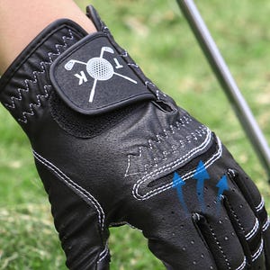May include: Black golf glove with white stitching and a golf ball logo. The glove has a breathable design with small perforations and blue arrows indicating airflow. The wrist strap features a logo with a golf ball and crossed clubs.