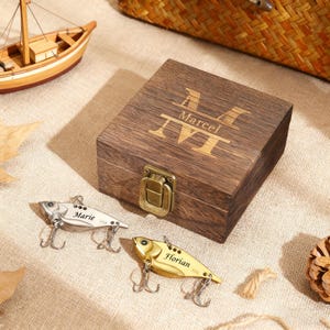 May include: A dark wooden box with the name "Marcel" engraved on the lid, alongside two fishing lures. One lure is silver, the other gold, each with a name. A small wooden boat is in the background.