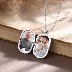 May include: A silver-toned oval locket necklace featuring two internal photo compartments. The left side displays a portrait of an older man in a grey sweater, while the right side shows a couple embracing. The locket hangs from a delicate chain.