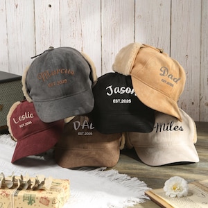 Könnte beinhalten: Eine Auswahl an personalisierten Trapper-Mützen in Grau, Schwarz, Burgunderrot und Beige. Jede Mütze zeigt einen gestickten Namen, wie "Marcus" und "Jason", mit "EST. 2025" darunter. Die Mützen haben Ohrenklappen und ein weiches Futter.