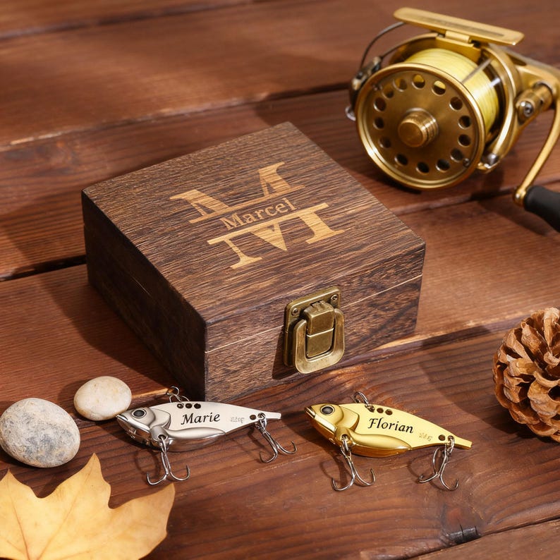 May include: A wooden box with the name "Marcel" engraved on the lid, next to a gold fishing reel and two fishing lures. One lure is silver and says "Marie", the other is gold and says "Florian". The lures have three hooks each.