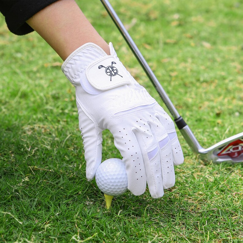 May include: White golf glove with a logo on the wrist, holding a golf ball on a tee. The glove has perforations for breathability and is on a green grassy surface. A golf club is in the background.