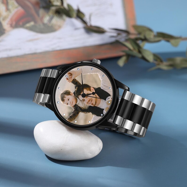 May include: A black wristwatch featuring a photograph of a man and child on the watch face. The watch has a black strap with silver detailing and is displayed on a blue surface next to a white stone.