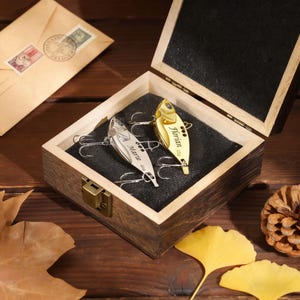 May include: A wooden box containing two fishing lures. One is gold with the name "Florian", the other silver with "Marie". The box is open, revealing a black interior, and rests on a wooden surface. Autumn leaves are scattered around.