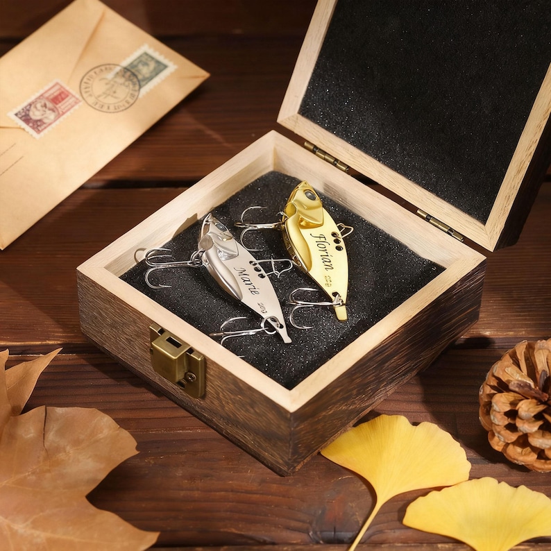May include: A wooden box containing two fishing lures. One is gold with the name "Florian", the other silver with "Marie". The box is open, revealing a black interior, and rests on a wooden surface. Autumn leaves are scattered around.