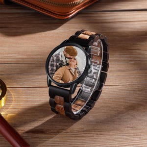 May include: A black wristwatch with a wooden strap and a round face. The watch face features a colour photograph of a couple. The strap combines dark wood and lighter wood tones. The watch is displayed on a wooden surface.