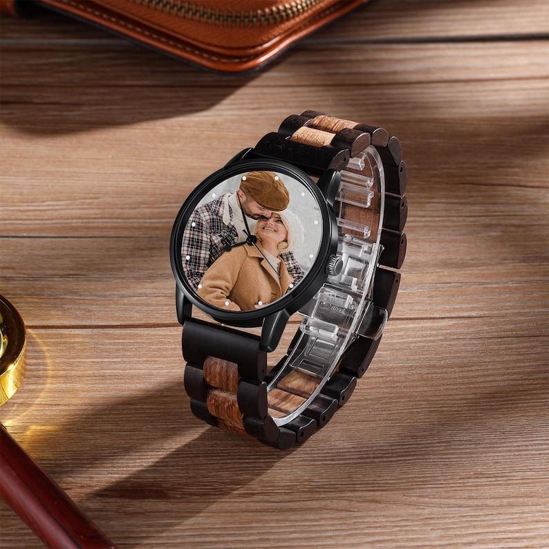 May include: A black wristwatch with a wooden strap and a round face. The watch face features a colour photograph of a couple. The strap combines dark wood and lighter wood tones. The watch is displayed on a wooden surface.