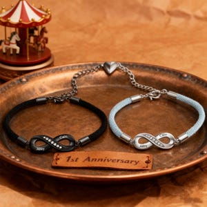 Op de afbeelding: Twee bijpassende armbanden met een oneindigheidssymbool, één zwart en één lichtblauw, gepresenteerd op een koperkleurig dienblad. De zwarte armband heeft de namen "Abby" en "Cain" gegraveerd. De lichtblauwe armband heeft dezelfde namen. Een klein houten bordje leest "1st Anniversary".