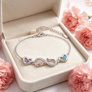 Könnte beinhalten: Silbernes Armband mit Unendlichkeitszeichen und dem Wort "Mom". Das Armband hat herzförmige Akzente mit lila und blauen Edelsteinen. Präsentiert in einer weißen Schmuckschatulle, umgeben von rosa Blumen. Ein schönes Geschenk.