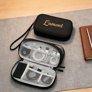 Puede incluir: Organizador de viaje negro con el texto dorado "Lamont". El organizador abierto revela compartimentos con cables de carga blancos, un teléfono negro y otros accesorios tecnológicos. También se ven un cuaderno y un bolígrafo marrones.
