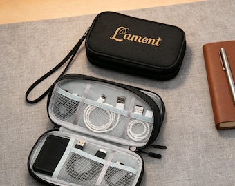 Può includere: Organizer da viaggio nero con il testo dorato "Lamont". L'organizer aperto rivela scomparti con cavi di ricarica bianchi, un telefono nero e altri accessori tecnologici. Sono visibili anche un taccuino e una penna marroni.