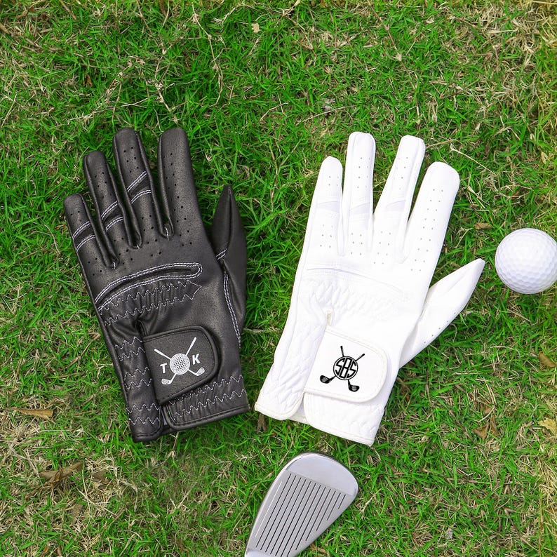 May include: Black and white golf gloves with a golf ball and club on green grass. The black glove has white stitching and the initials T and K. The white glove has the initials SBS. A golf club is also visible.