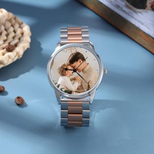 May include: A silver and rose gold wristwatch featuring a photograph of a father and child on the watch face. The watch has a round face and a stainless steel band with rose gold accents. The watch is displayed on a light blue surface.