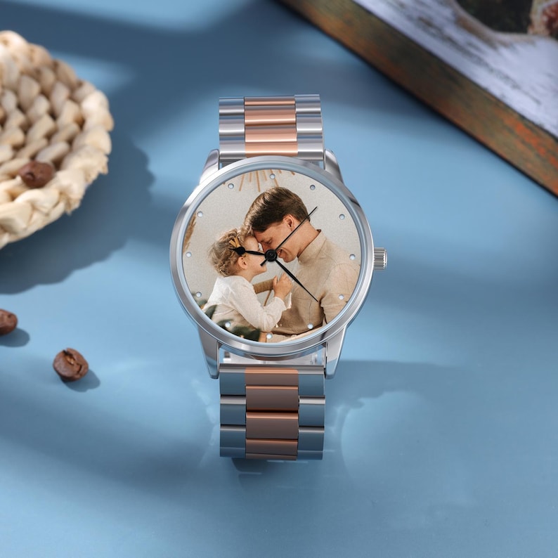 May include: A silver and rose gold wristwatch featuring a photograph of a father and child on the watch face. The watch has a round face and a stainless steel band with rose gold accents. The watch is displayed on a light blue surface.
