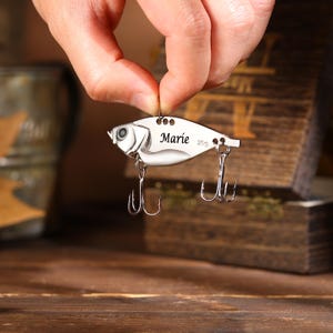 May include: A silver fishing lure in the shape of a fish, with two treble hooks and the name "Marie" engraved on it. The lure weighs 20g. The lure is held up by a person's hand.
