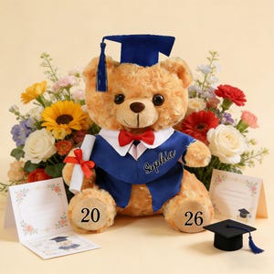 May include: A plush teddy bear wearing a blue graduation gown and cap, with a red bow tie. The bear holds a diploma tied with a red ribbon. The name "Sophia" is embroidered on the gown. Numbers "20" and "26" are on the bear's paws. A small graduation cap and cards are also present.