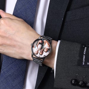 May include: Black wristwatch featuring a family photo on the watch face. The watch has a black metal strap and a round dial. The watch is worn on a wrist, with a dark suit and tie visible.