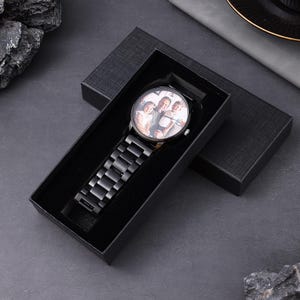 May include: A black wristwatch with a metal strap and a round face displaying a family photograph. The watch is presented in a black gift box, set against a dark grey background. The watch strap is made of linked metal pieces.