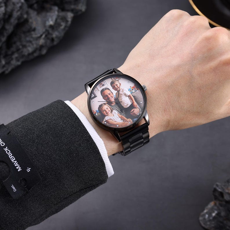 May include: Black wristwatch featuring a family photo on the watch face. The watch has a black metal strap and a circular dial. The watch is worn on a wrist, with a dark suit jacket visible in the background.