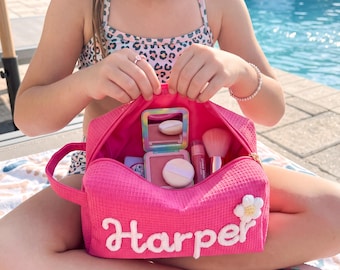 Puede incluir: Un neceser de color rosa brillante con textura de gofre, el nombre Harper en letras cursivas blancas y una aplicación de flor blanca. El neceser abierto contiene un espejo compacto, un colorete con borla, brillo labial y una brocha.