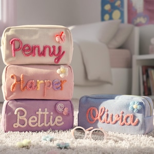 May include: A stack of four corduroy cosmetic bags in pastel colors: cream, pink, lavender, and blue. Each bag has a name embroidered on it: Penny, Harper, Bettie, and Olivia. Decorative floral and butterfly accents adorn the bags. Sunglasses and small flower-shaped accessories are in front.
