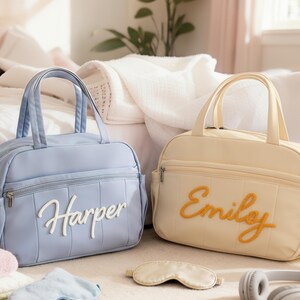Puede incluir: Dos bolsas de beb&eacute; personalizadas, una azul claro con "Harper" en escritura blanca y la otra color crema con "Emily" en escritura naranja. Una m&aacute;scara para dormir y auriculares est&aacute;n cerca. Las bolsas tienen asas superiores y bolsillos laterales.