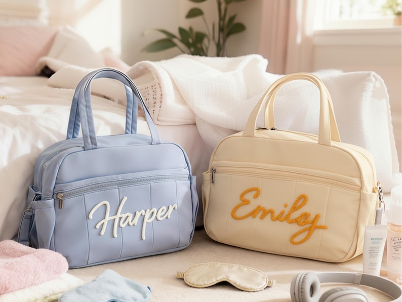 Puede incluir: Dos bolsas de beb&eacute; personalizadas, una azul claro con "Harper" en escritura blanca y la otra color crema con "Emily" en escritura naranja. Una m&aacute;scara para dormir y auriculares est&aacute;n cerca. Las bolsas tienen asas superiores y bolsillos laterales.